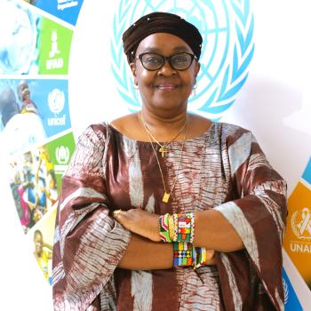 man in brown dress and head piece stands in front of SDG symbol 