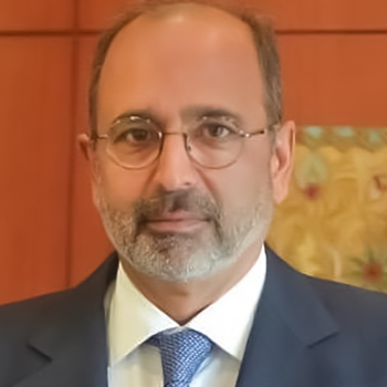 A man in a grey suit and blue tie and glasses poses for the camera next to a blue UN flag.