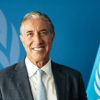 ASG looks at the camera smiling with the UN flag by his side and the UN logo in the background