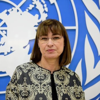 A woman wearing a black and gold jacket stands in front of the UN logo