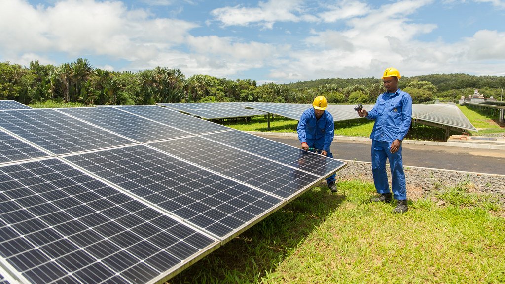 Two workers in blue coveralls and yellow hard hats inspect rows of solar panels on a grassy site, with trees and a cloudy blue sky behind them.