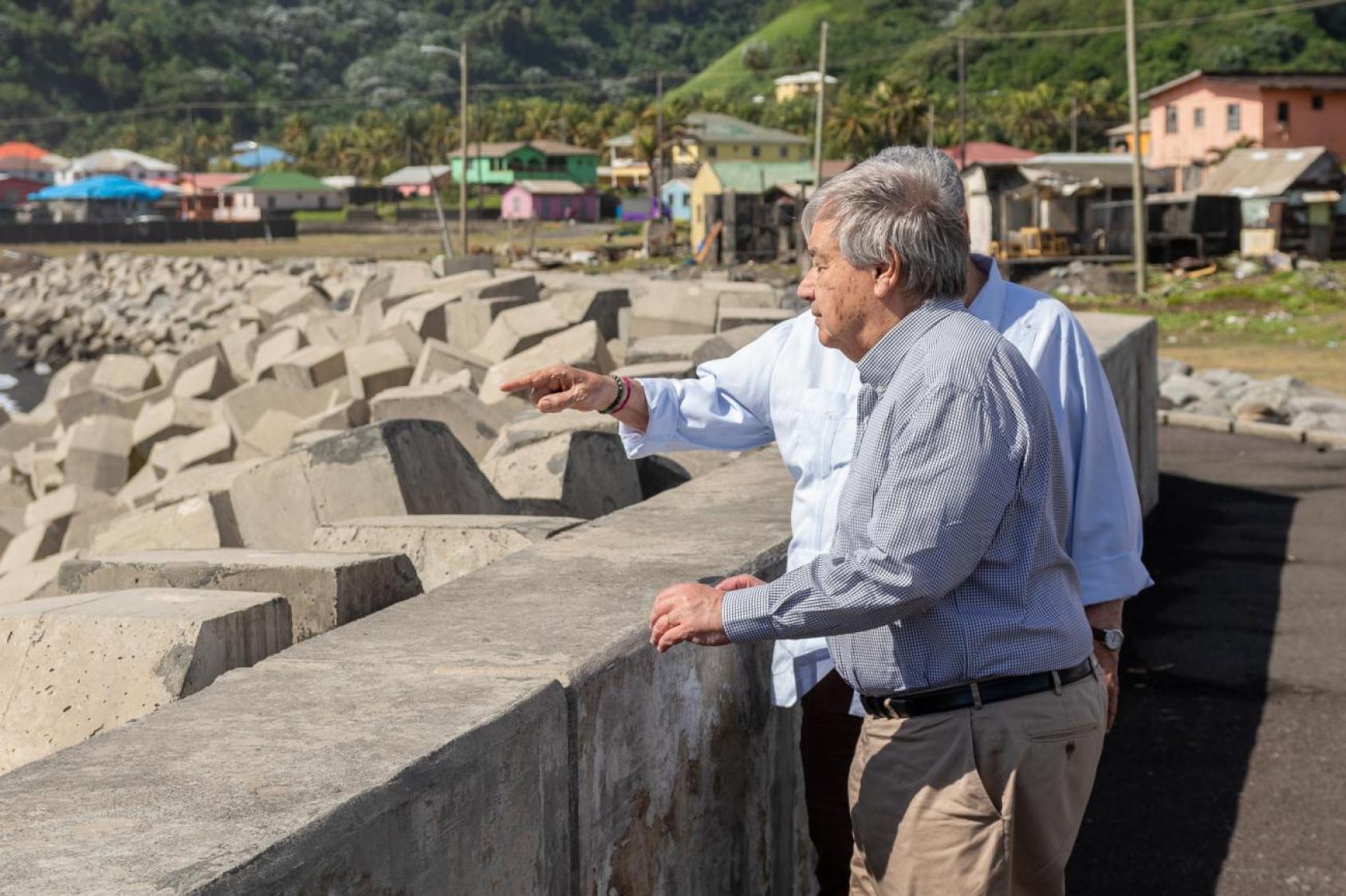 Two men, in blue shirts and beige pants, stand at a short wall overlooking the sea.