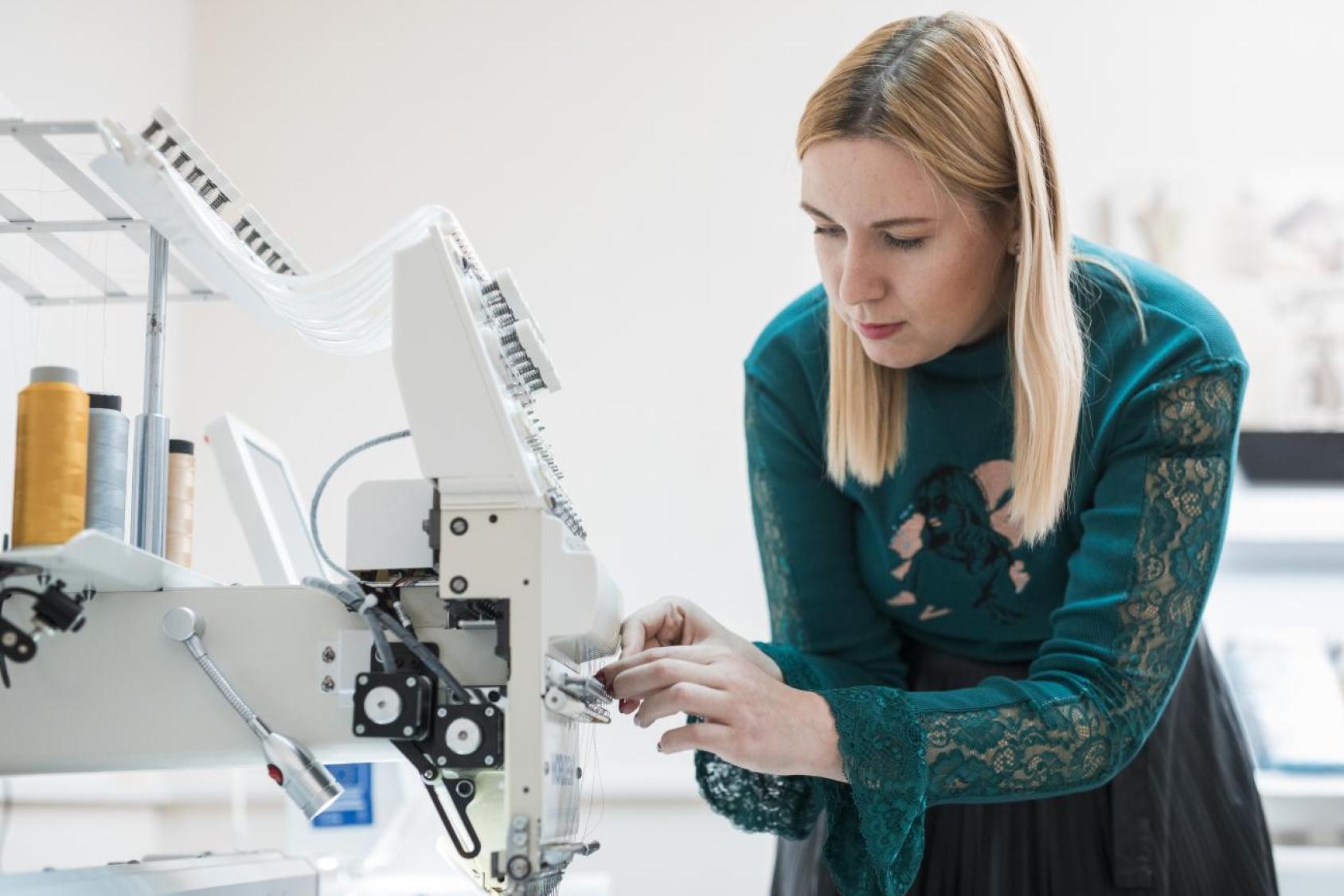 Person in a teal dress with lace detailing operating a sewing machine, with spools of thread visible