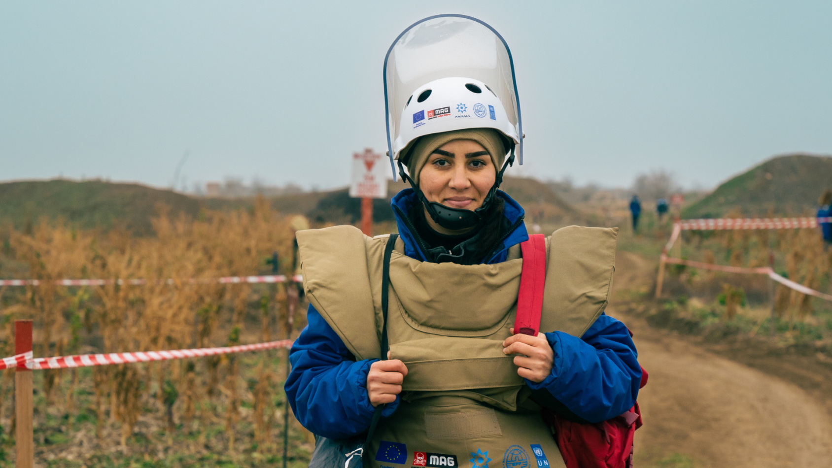 A woman wearing demining clothing, a helmet and chest guard, looks at the camera