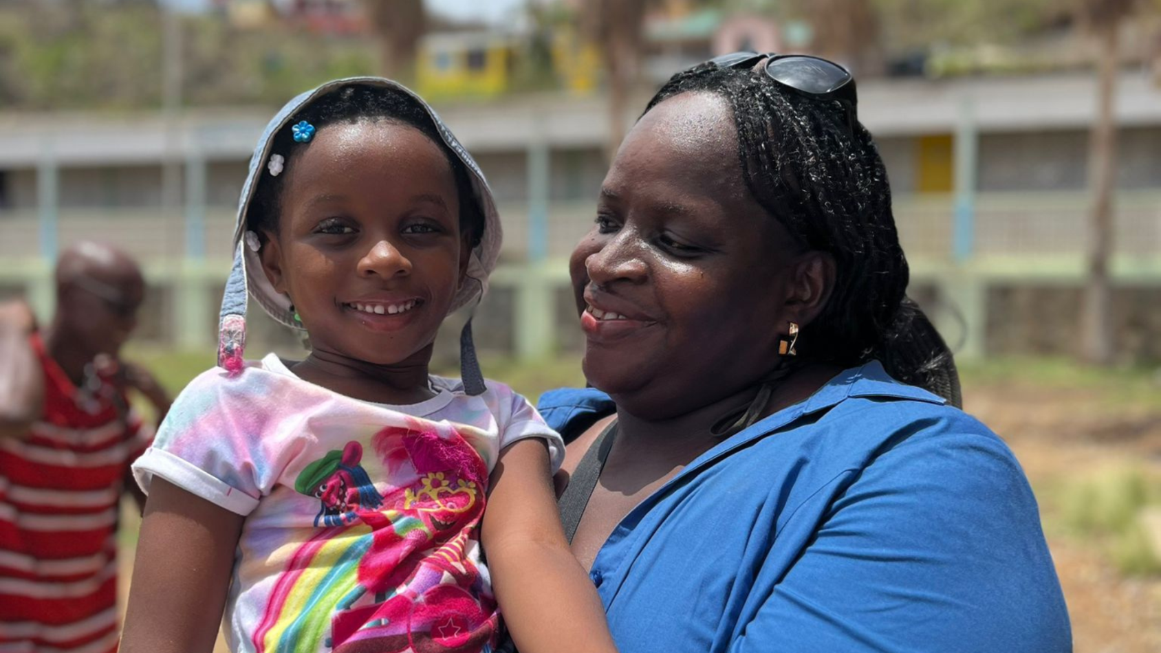 A woman is holding a child and standing outdoors, they are both looking at the camera and smiling 