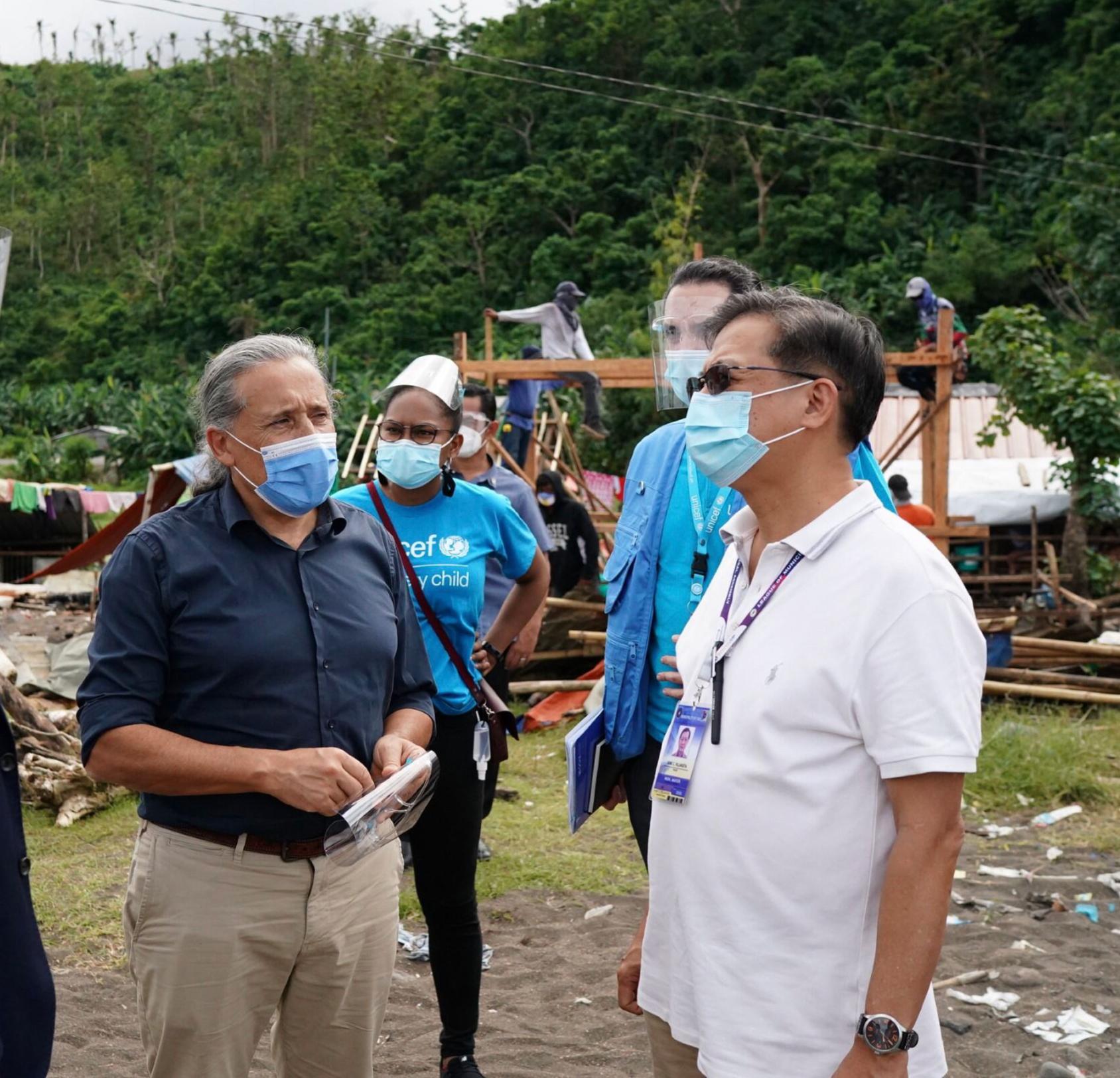 Two men wearing face masks are in an outdoor setting and engaging in a discussion there are two others in the background