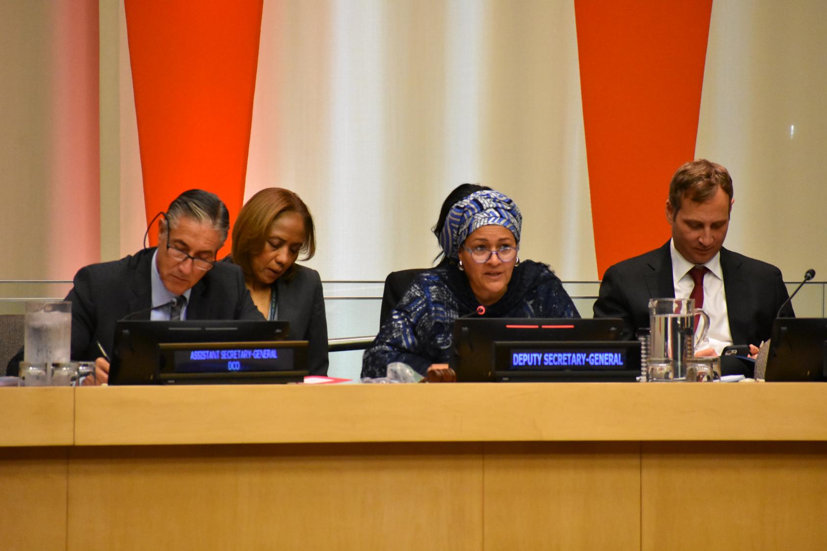 A woman and man are seated at a podium in the foreground, three others are seated behind them. They are seated at the head table of a conference room