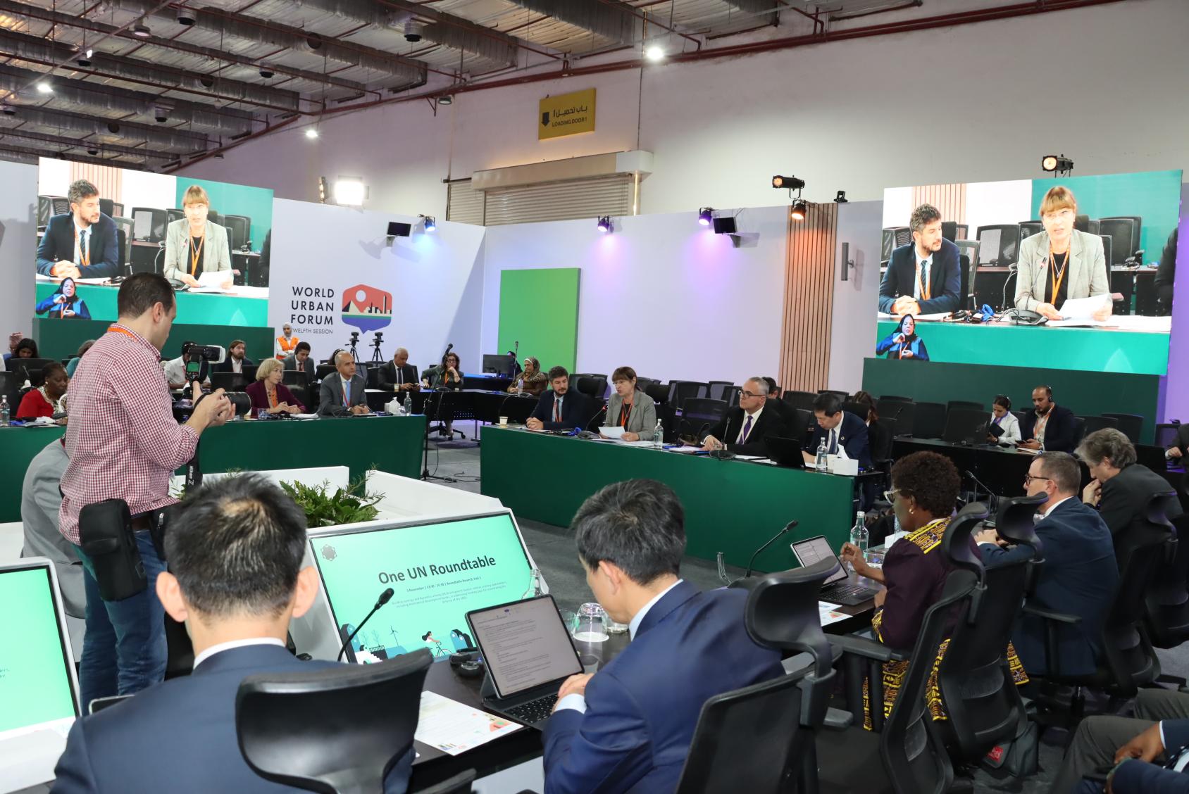 A group of people in a conference room, there are banners with the logo of the World Urban Forum and the One UN Round Table