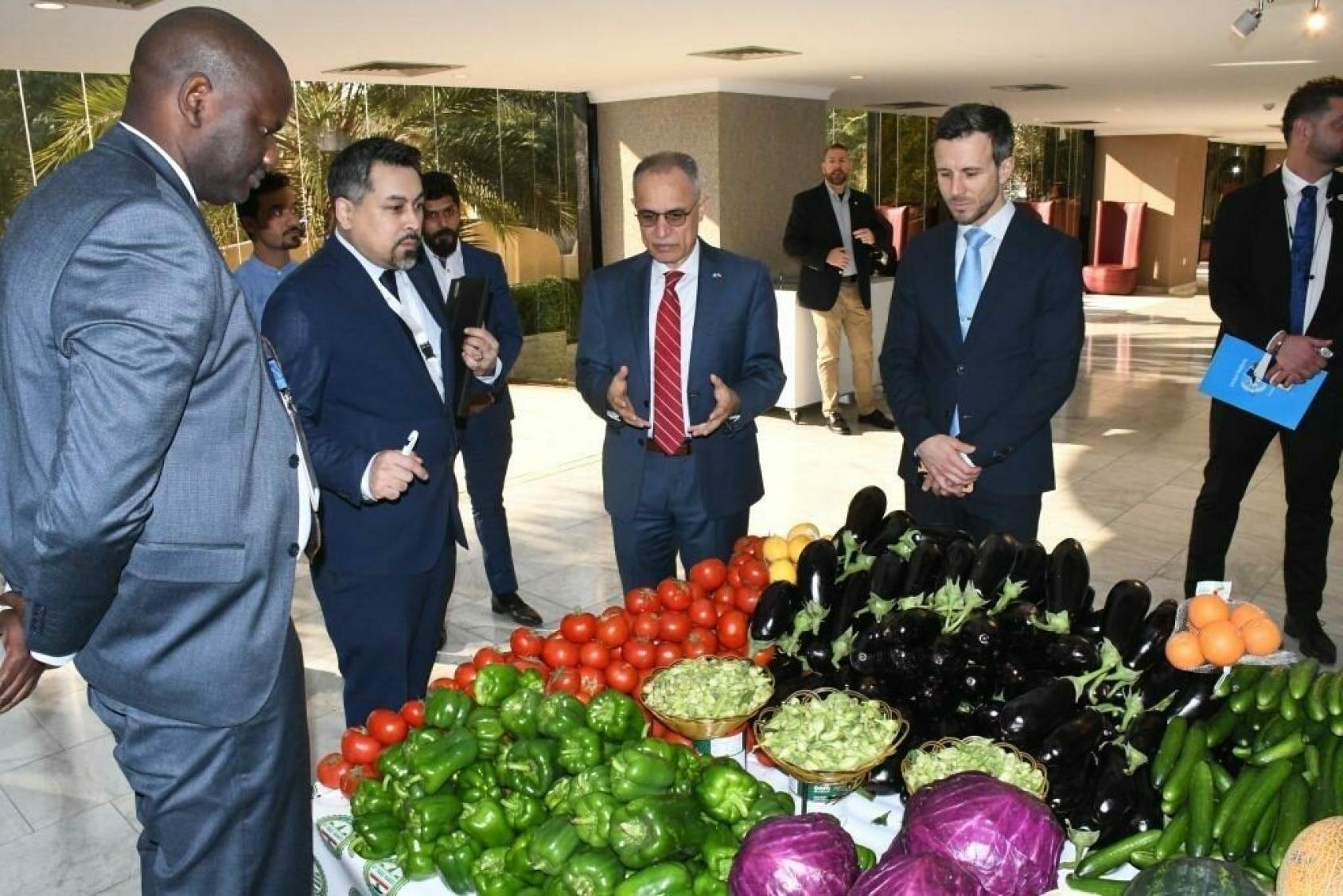 A group of people in an indoor setting wearing formal attire, having a discussion around a display of produce