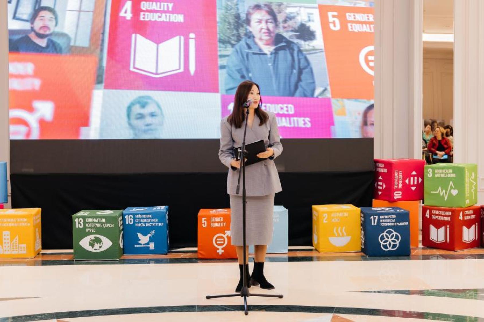 A woman stands at a microphone in front of Sustainable Development Goals banners and cube displays, advocating for disability inclusion.