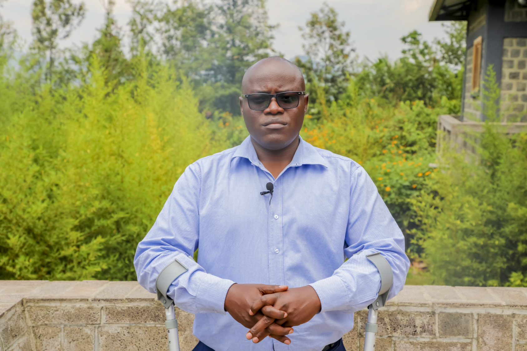 A man in blue shirt and sunglasses standing in front of green and lush vegetation, portraying disability and inclusion