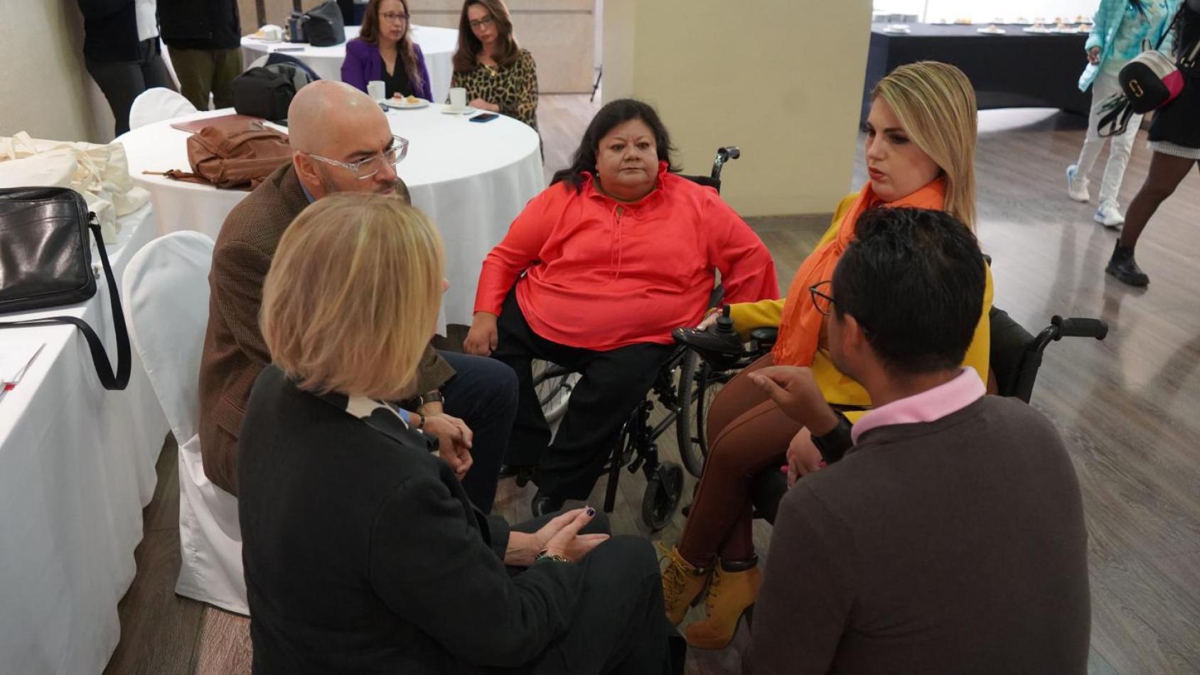 Diverse group of people, including a woman in a wheelchair, gathered around a table in a conference setting, showcasing disability representation and inclusion