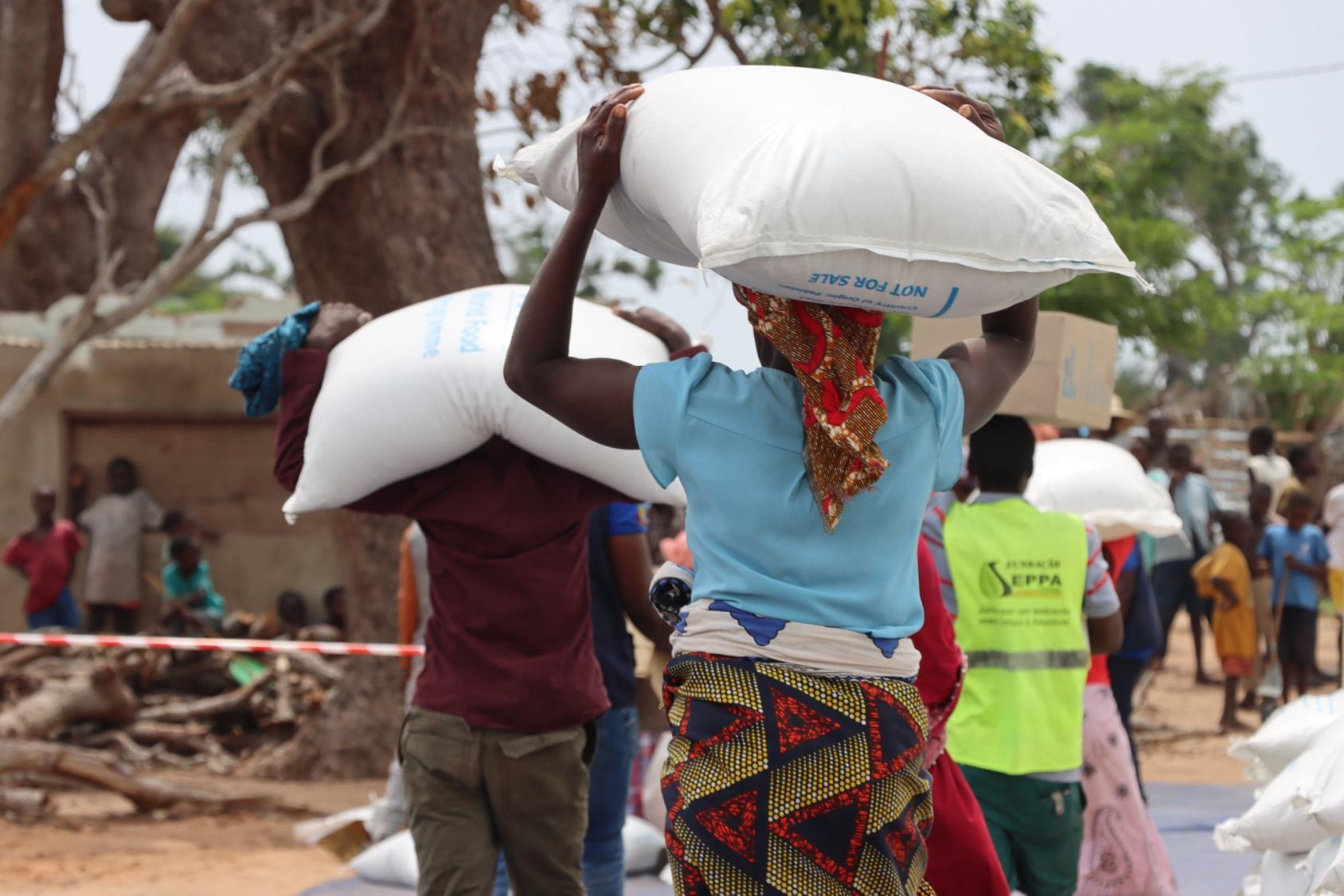 A woman is carrying a large bag of grain on her head, her back is turned to the camera