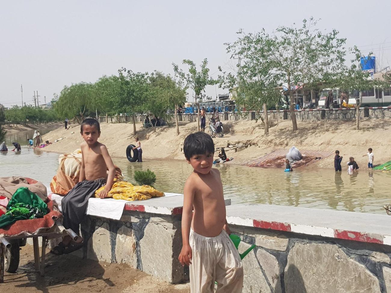 Children stand near a river with people washing clothes and carpets in a water canal.