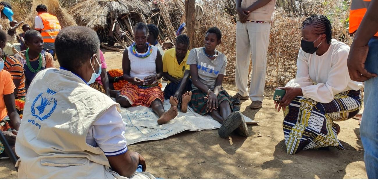 women kneels down speaking to a group of people on the ground 