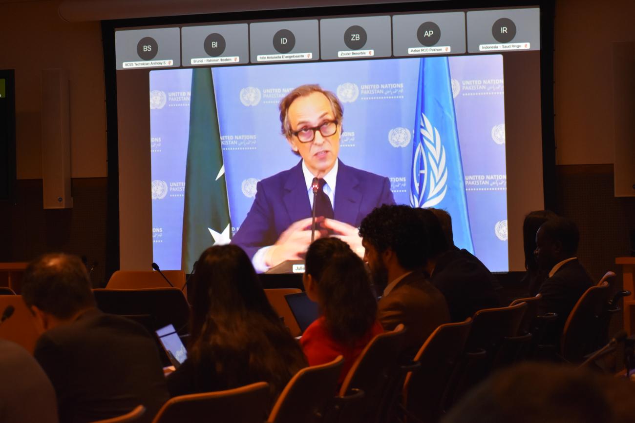 a man speaking on a screen to a meeting of people 