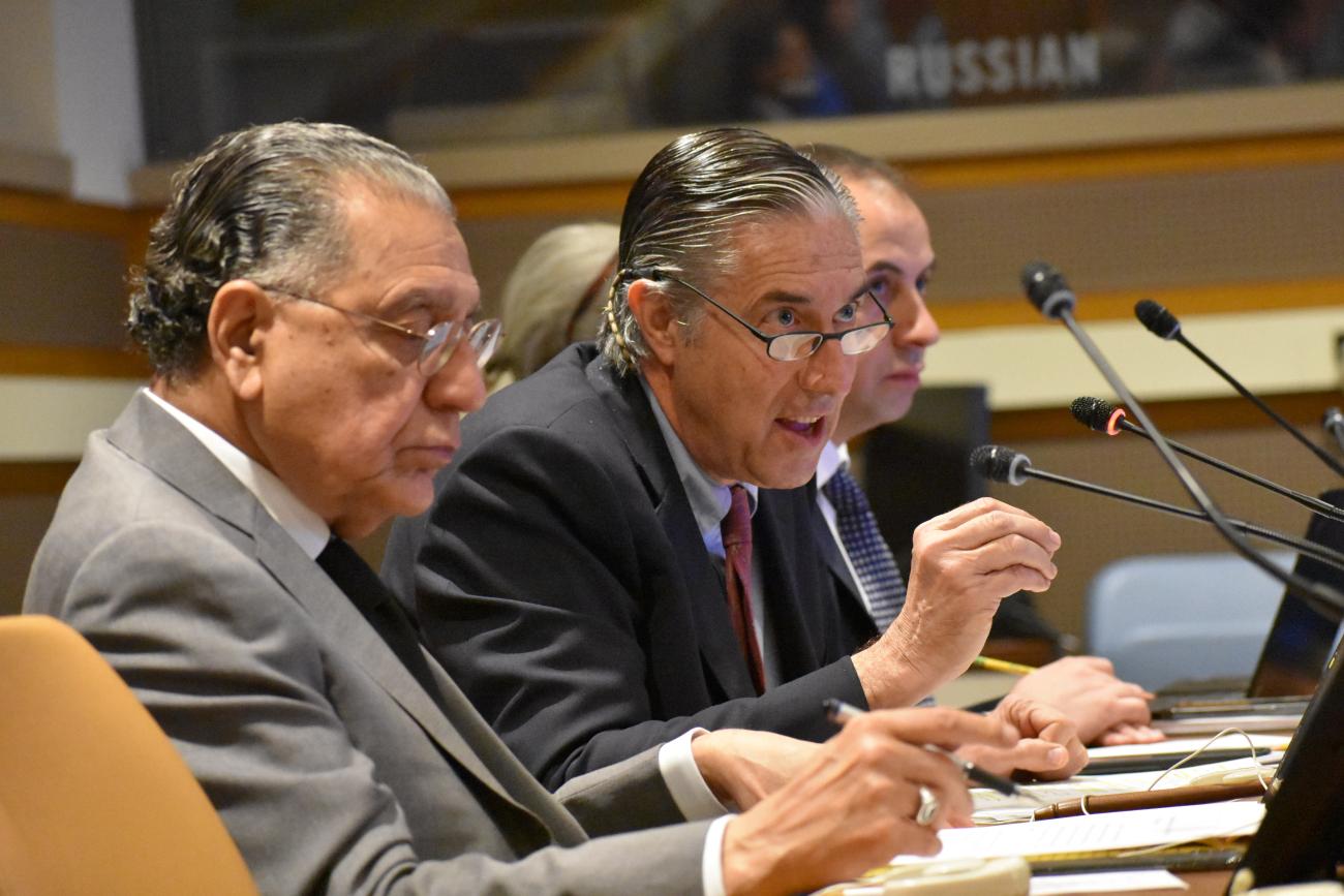 two men in grey suits speak into micro phones at a meeting