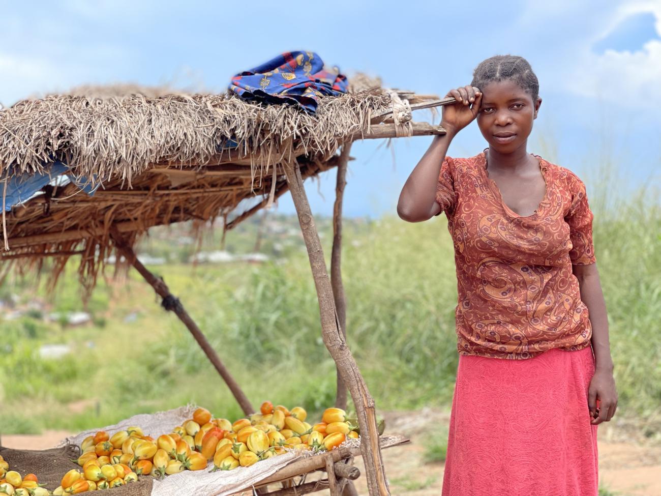 woman in orange top stands outside leaning against a straw roofed structure 