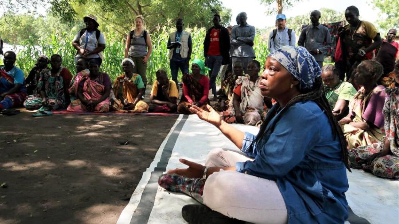 woman sits on the ground in blue tunic speaking to other women sitting in a circle 