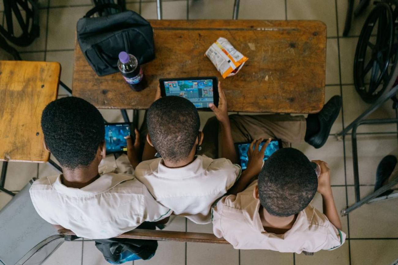 view from above of three school children sitting at a desk looking at a tablet