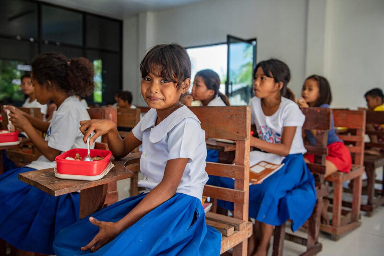 school children in blue and white uniform sit at desks in classroom