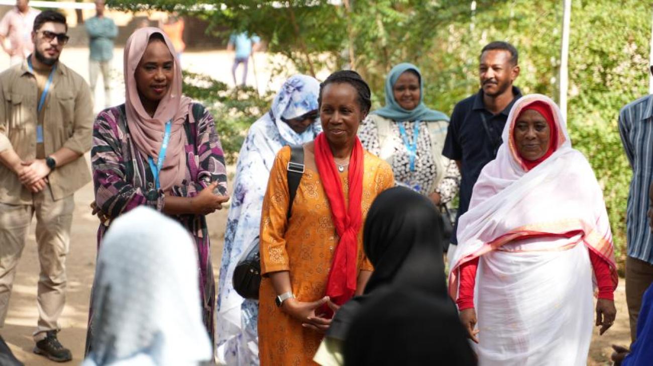 Women in tunics and free-flowing scarves stand in a semi-circle and talk, inside a larger circle of men and women.
