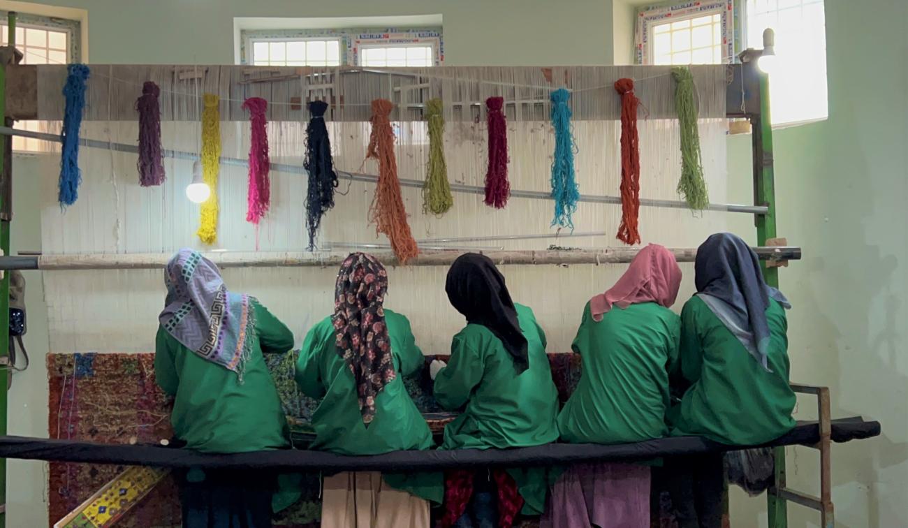 A group of Afghan women sitting on a loom, weaving a colorful rug. They are wearing traditional clothing and head coverings.