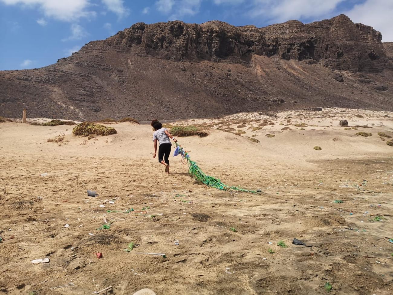 An individual dragging a large green net across a sandy area with sparse vegetation. In the background, there is a rugged mountain under a clear blue sky with few clouds. The ground is littered with various small pieces of debris.