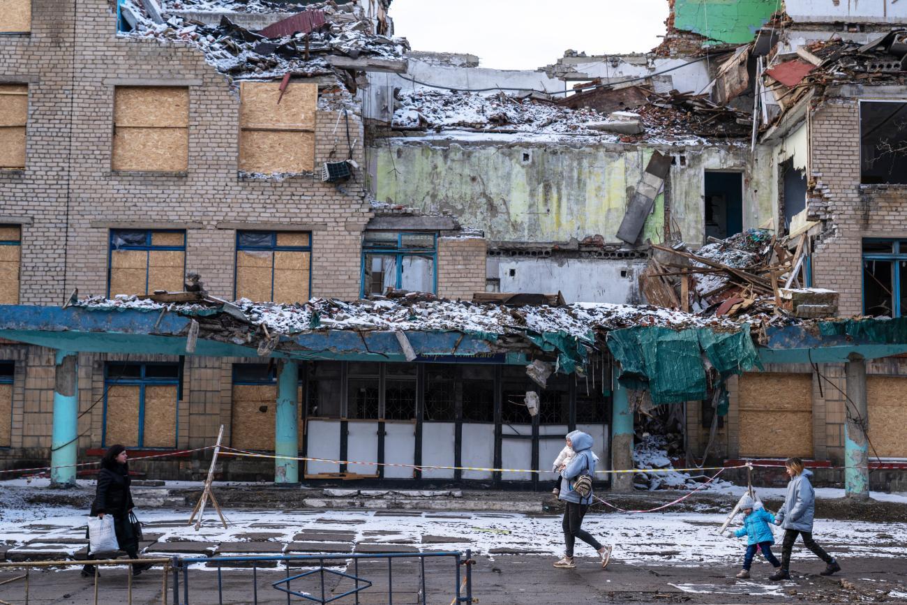 The image depicts a severely damaged multi-story building with its upper part largely destroyed, exposing the interior rooms. In the foreground, three people, including a child, walk past the building on snow-covered ground.