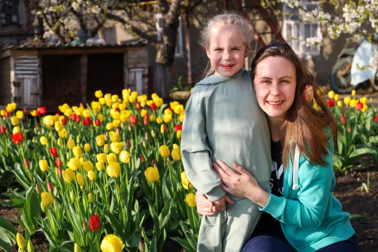 A woman in a green shirt hugs a child in a grey dress with blonde hair in front of a field of flowers