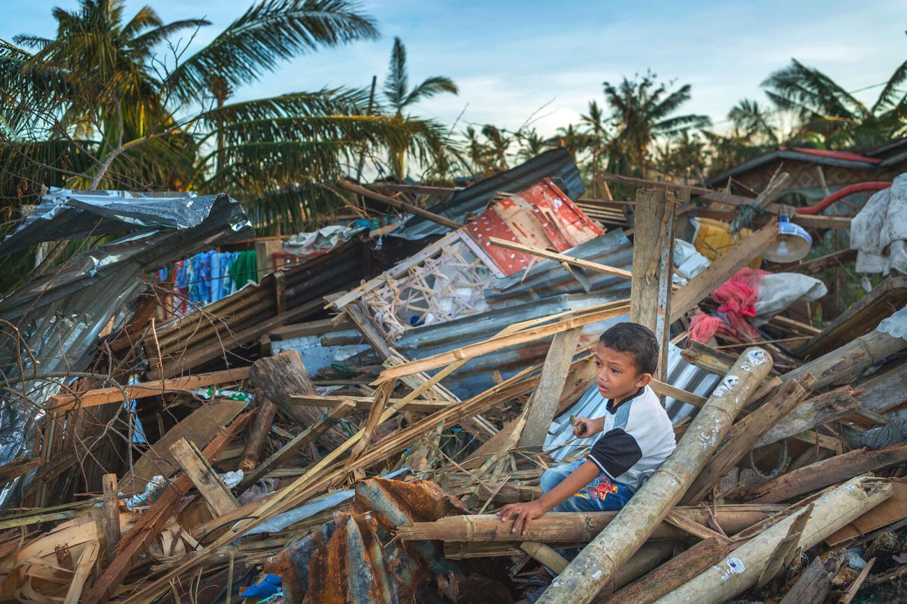 A collapsed house is in the background with a child seated on the rubble and crying