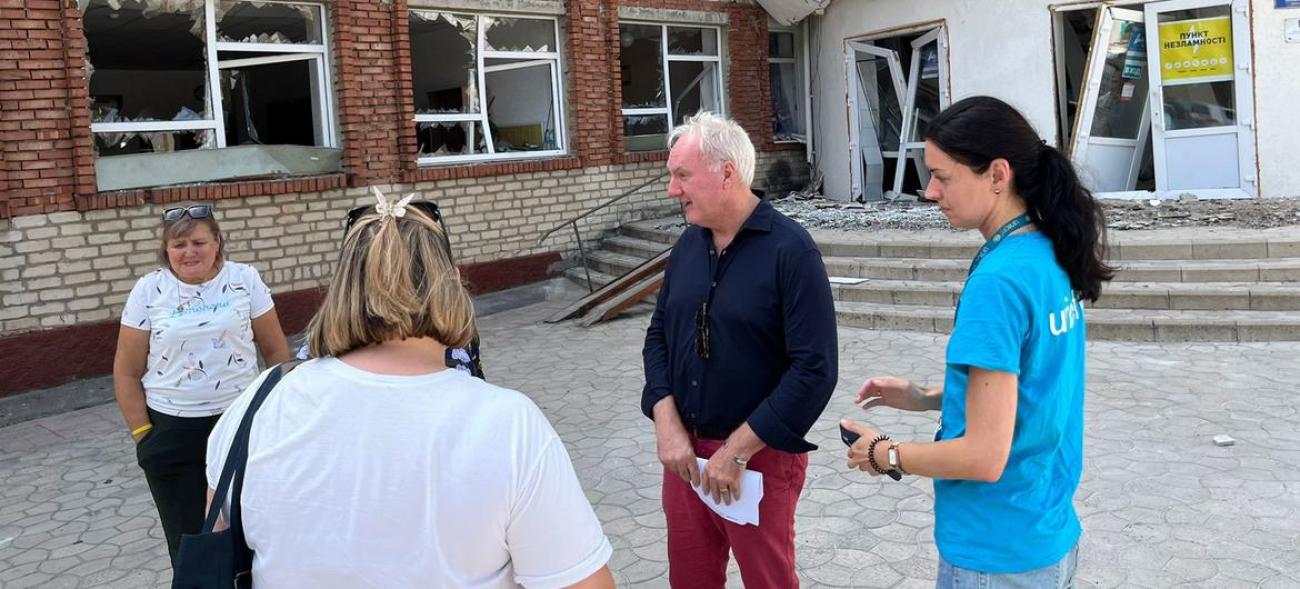 A man is wearing casual clothes and standing outdoors, he is surrounded by three women and stands in front of a partially destroyed building