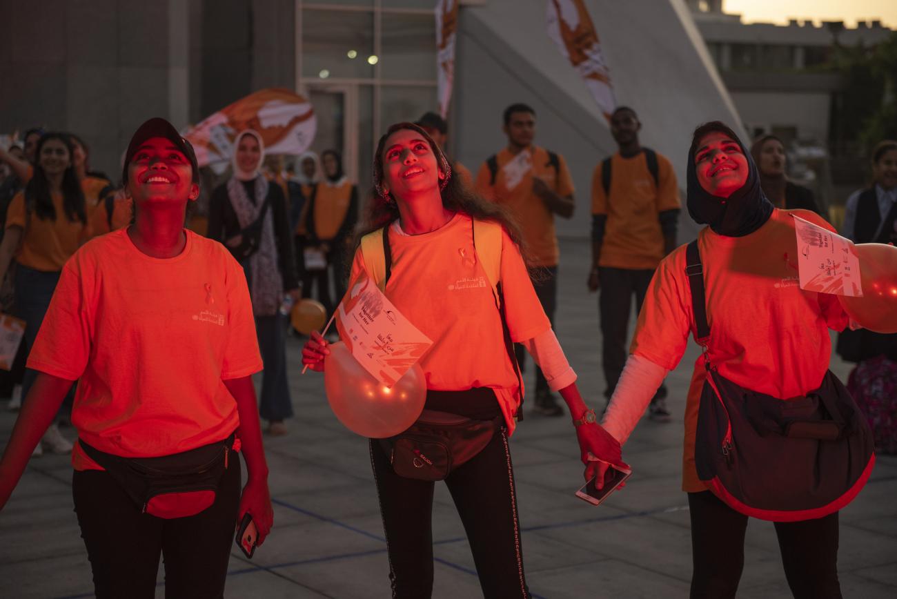 Three young girls are holding hands and looking up at the sky, they are wearing orange the world shirts marking the 16 days of activism against violence against women