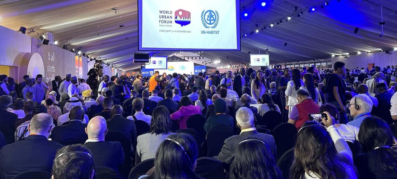 A large group of people in a conference room looking at a board that reads World Urban Forum 