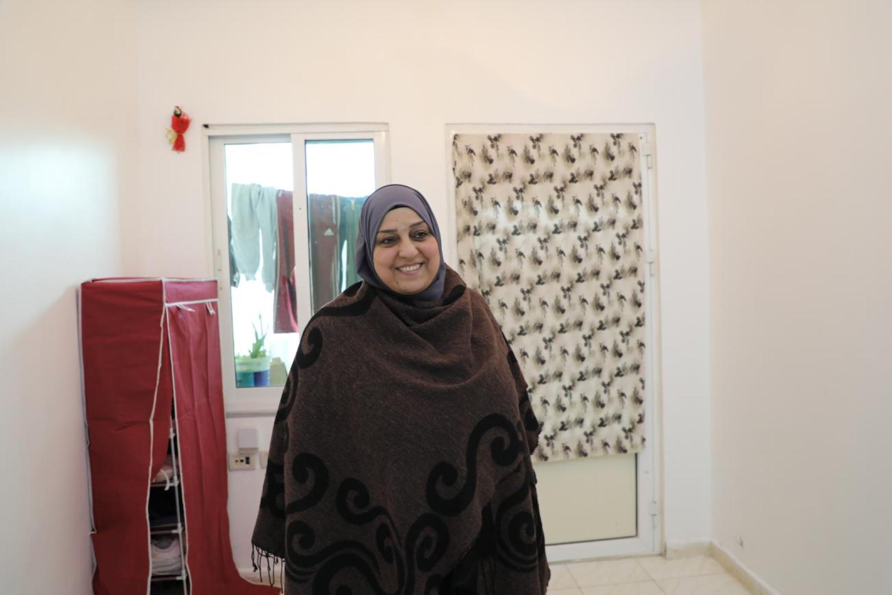 woman in dark headscarf stands in white room smiling at the camera