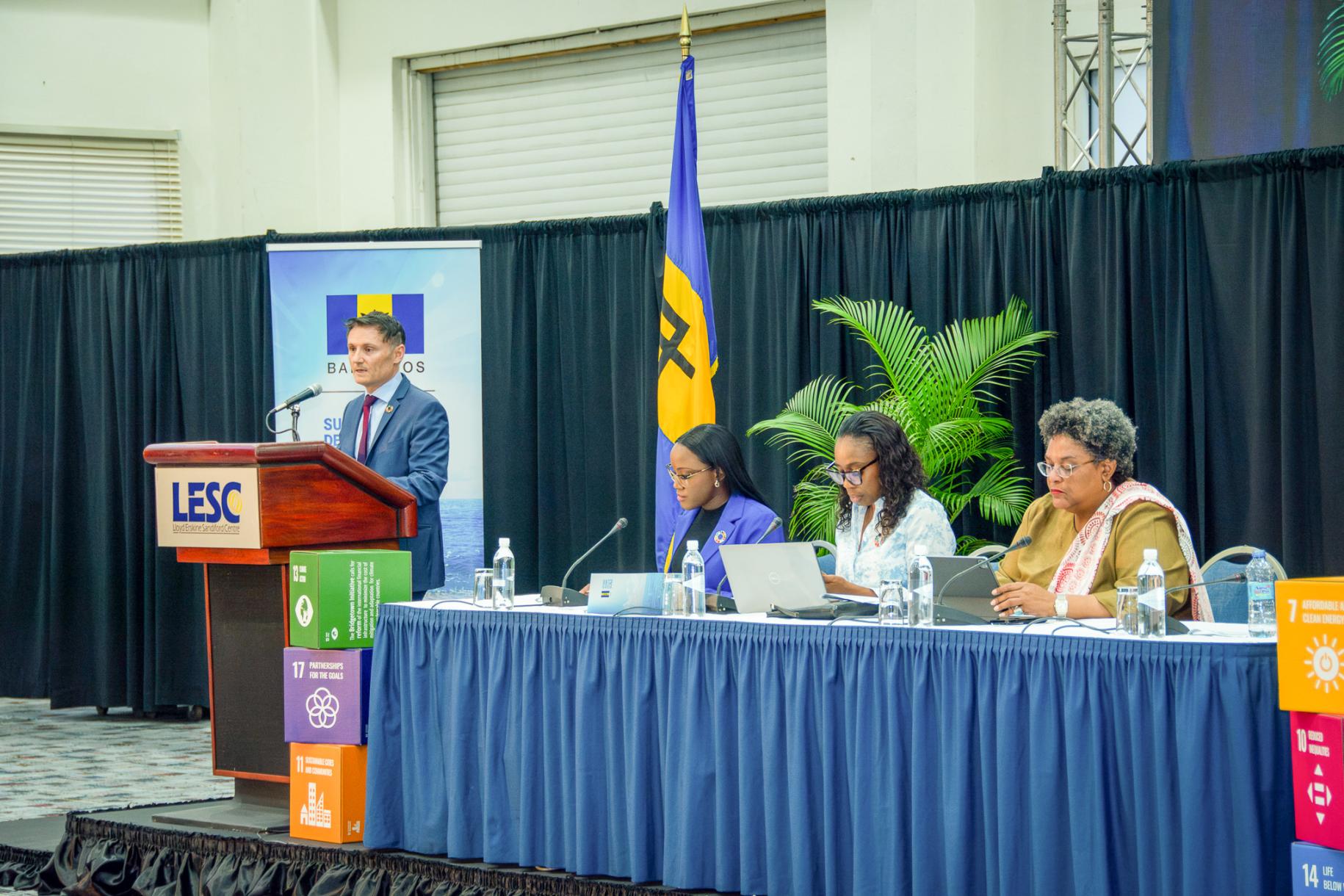 man in a suit speaks at podium next to panel of two women