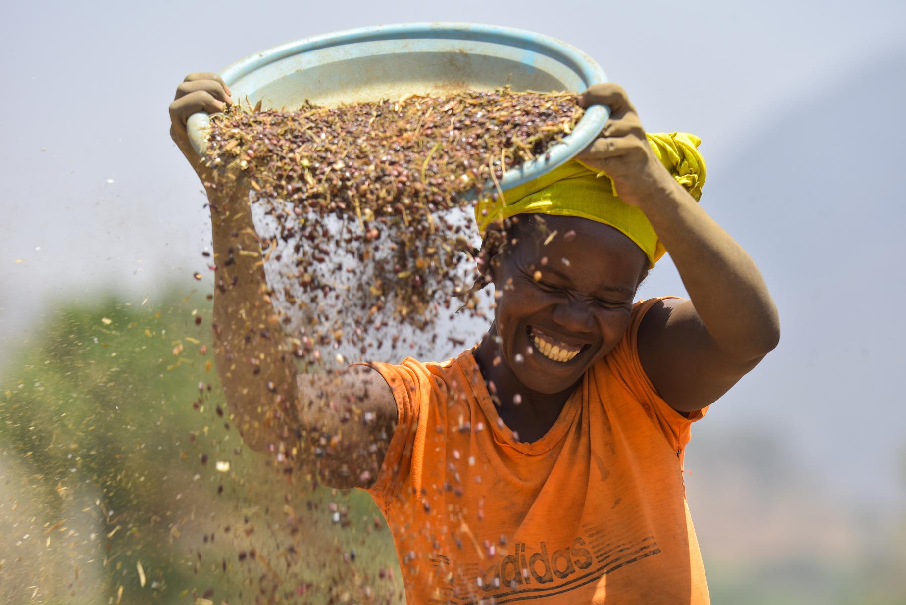 woman in orange top carries basket of grain above her head. 