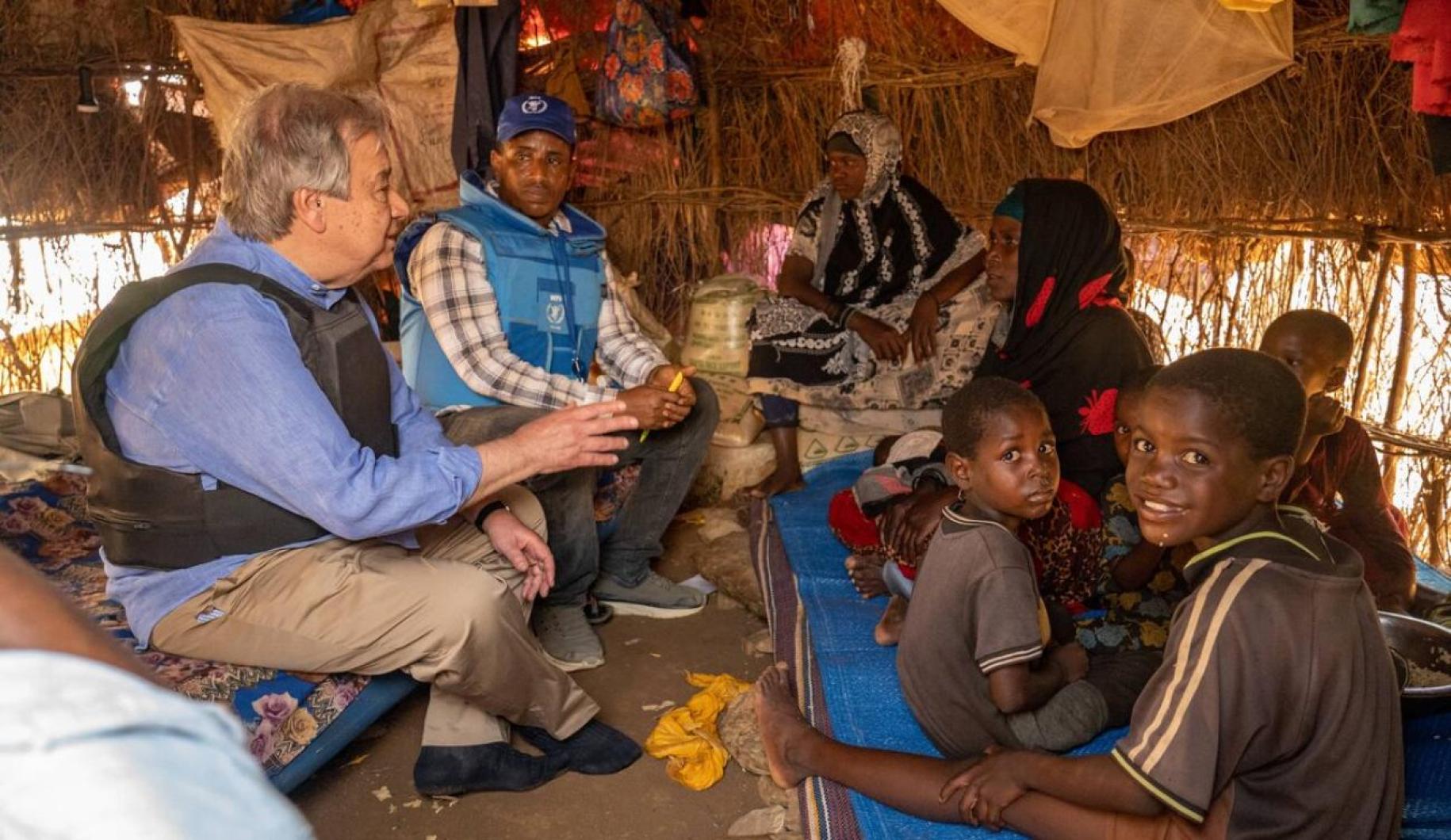 two men in bullet proof vests sit in hut and speak to a family with young children