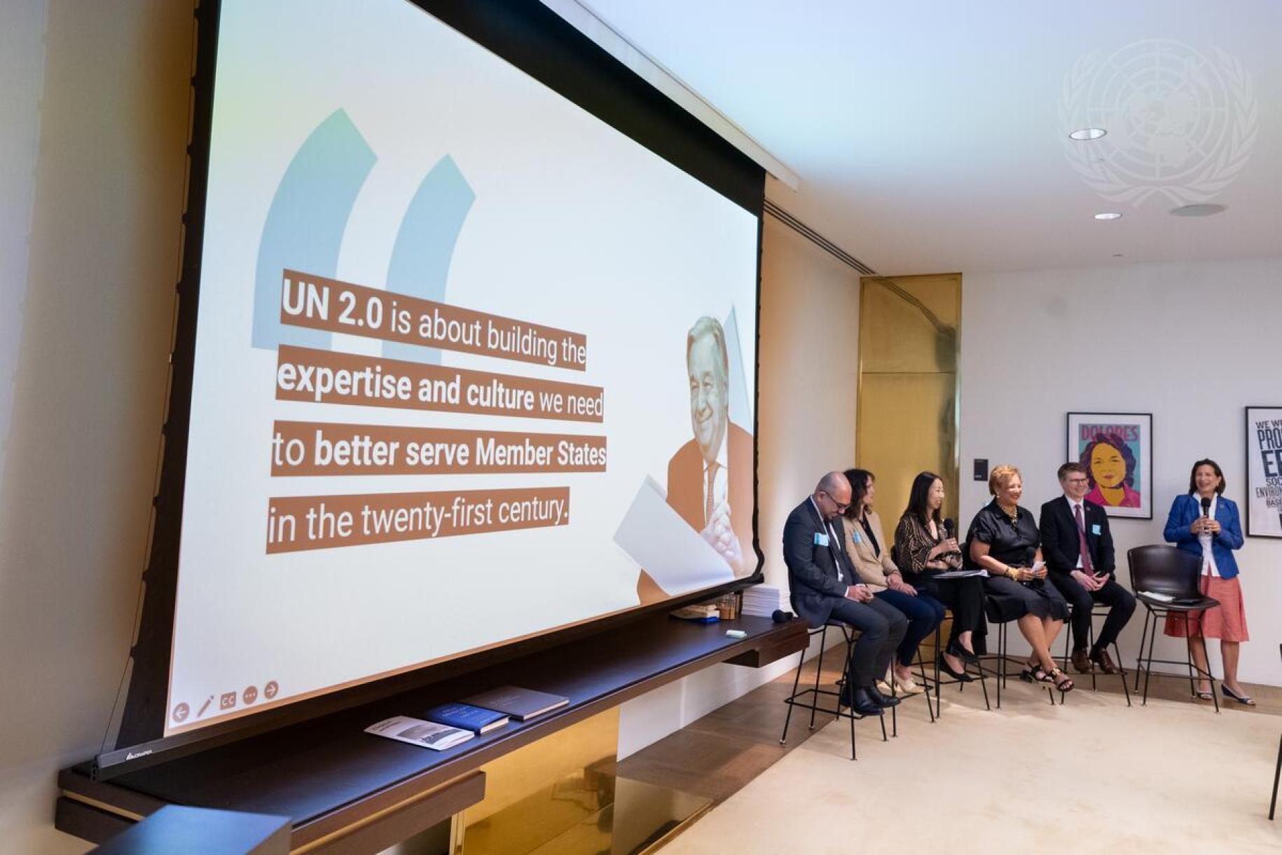 woman stands beside panel of people to present a powerpoint 