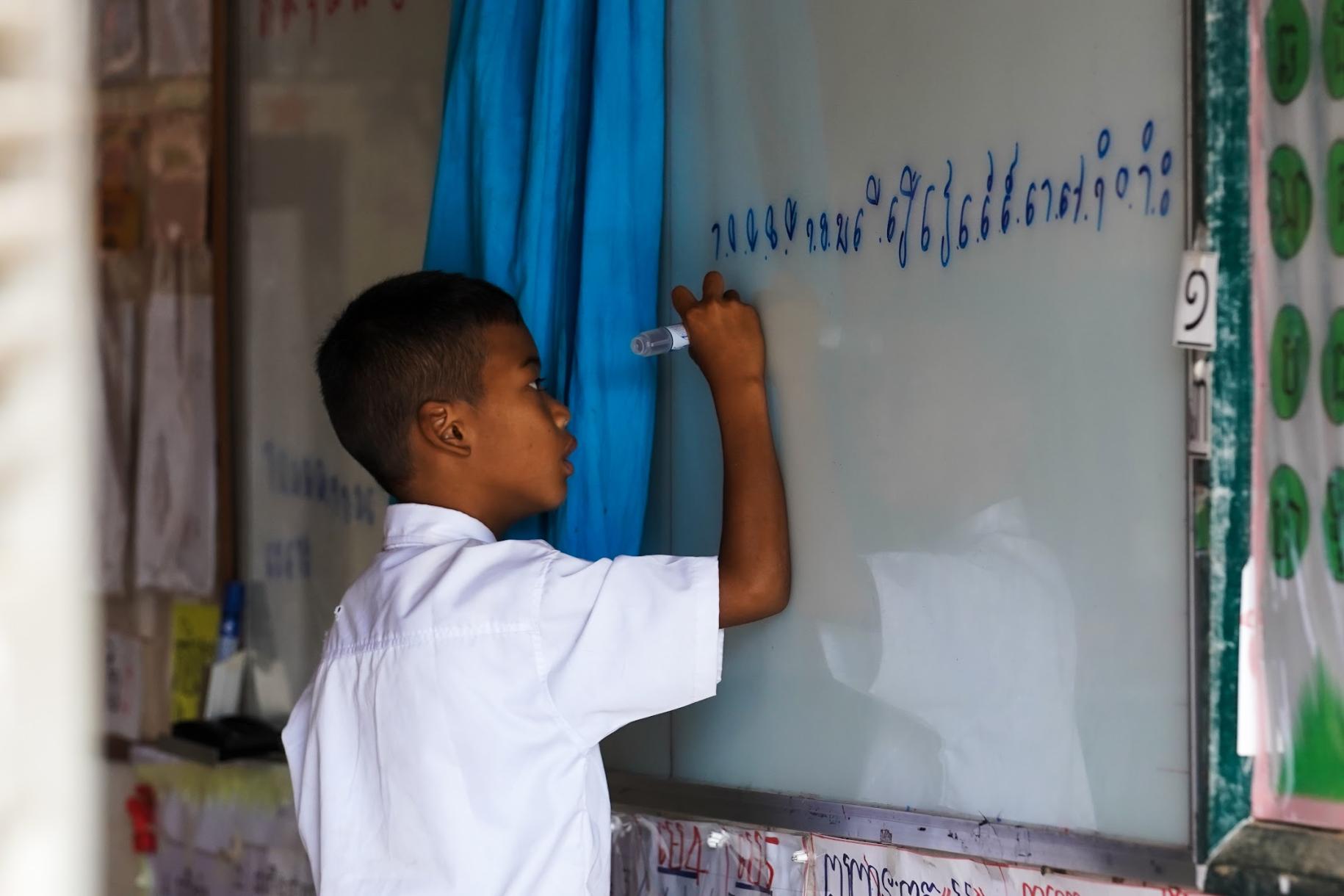 young boy in white top writes on white board in classroom