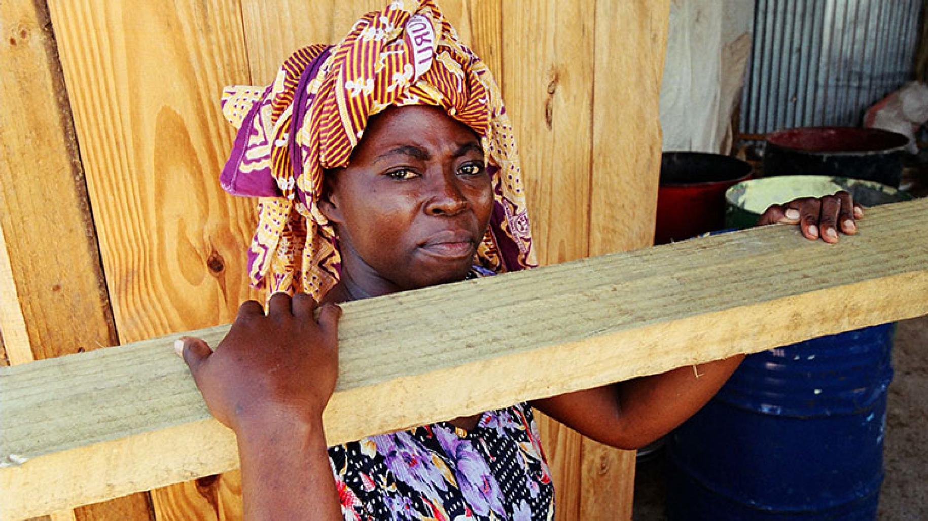 Women in colourful headscarf holds plank of wood on her shoulder and looks to the camera
