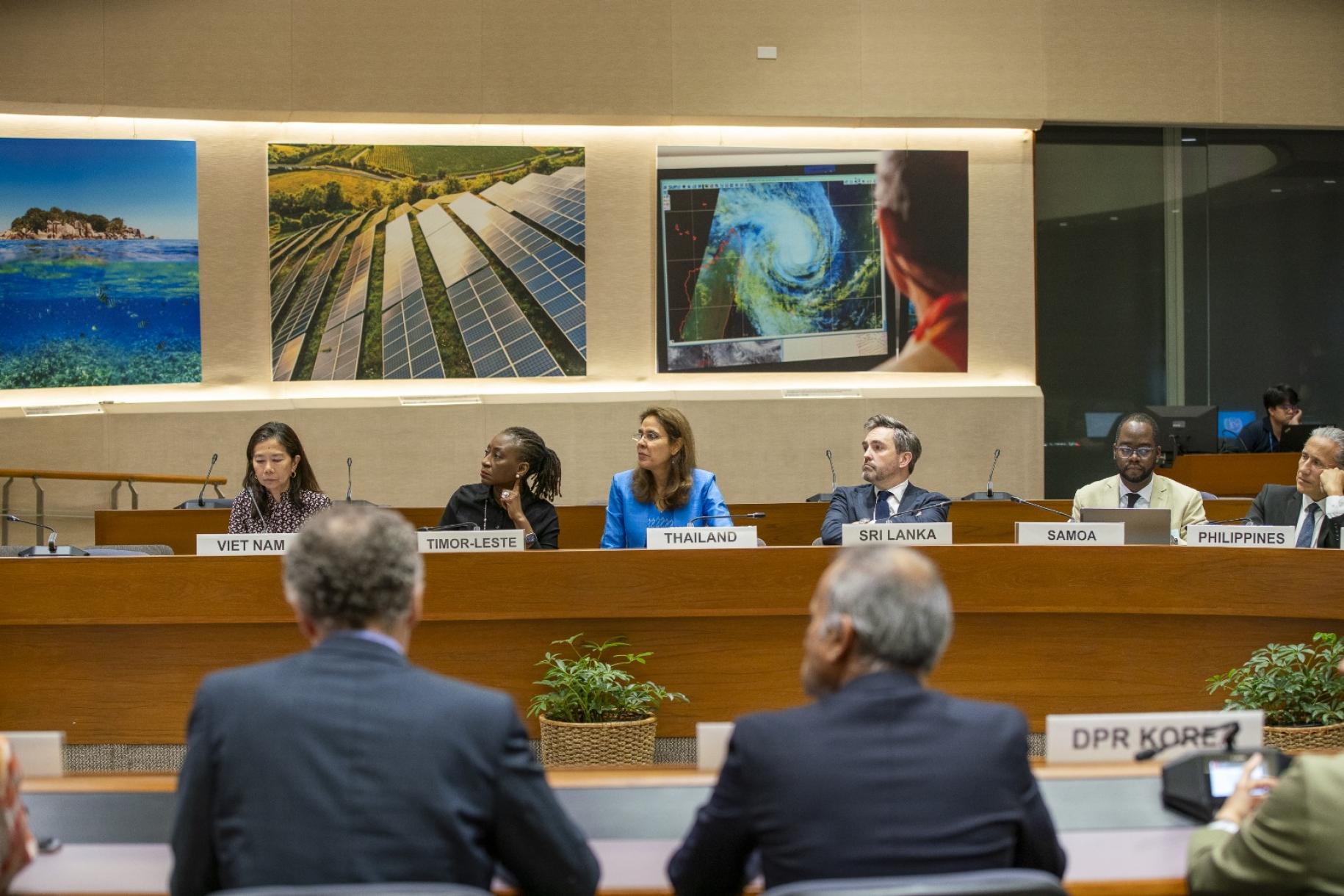 The image depicts a conference room with multiple individuals seated at a long, curved table. Each person is sitting behind a nameplate that indicates a country. 