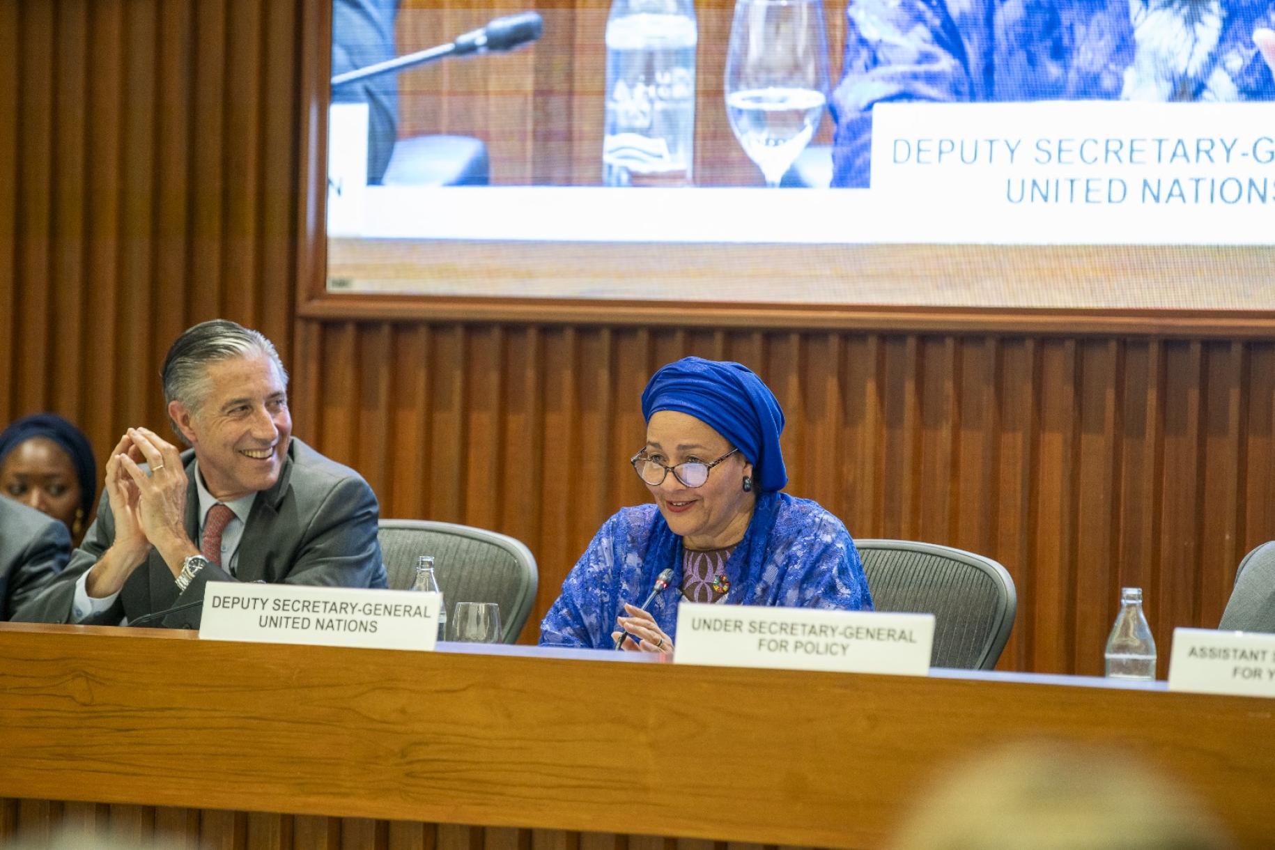 The image depicts a conference room with multiple individuals seated at a long, curved table. Each person is sitting behind a nameplate that indicates a country. 