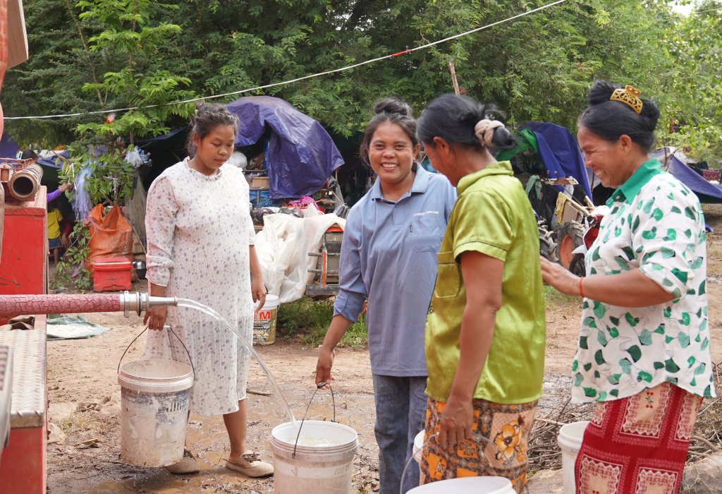 Four people stand outdoors near a temporary water setup, filling buckets from a pipe connected to a red machine. One person holds a bucket under a stream of water while others stand nearby with additional buckets. In the background, there are trees, tarps, and makeshift shelters with various items and equipment scattered around.