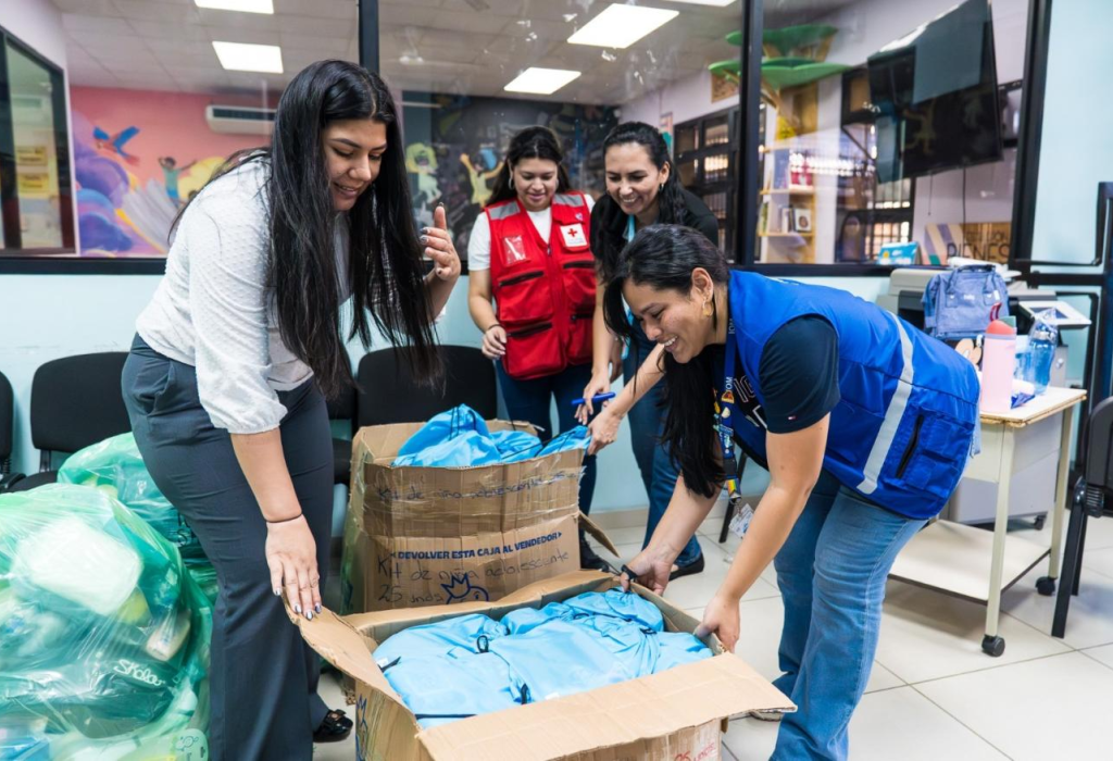 Several people in an indoor office setting unpack boxes of supplies, including blue bags, during a coordinated aid.