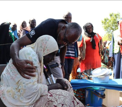 A group of people stand around as one person leans over to assist another seated individual during what appears to be an aid or community distribution event.